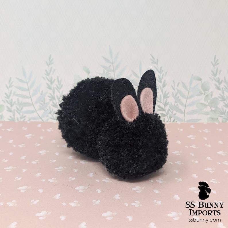 May include: Black yarn pom-pom bunny with pink felt ears. The bunny is lying on a pink background with white dots.