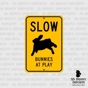 May include: Yellow road sign with black text and a black silhouette of a rabbit. The sign reads "Slow Bunnies at Play".