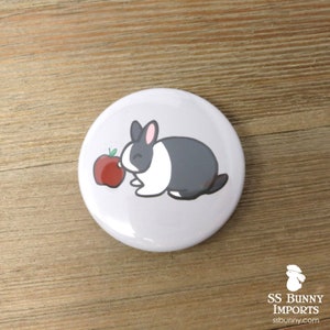 May include: A white and gray rabbit with black ears and eyes is sniffing a red apple. The rabbit is on a white button with a gray background.