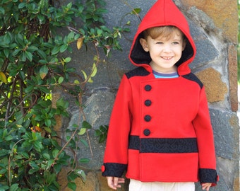 May include: A young child wearing a red hooded jacket with black trim and black buttons. The jacket has a decorative black band with a swirling pattern. The child is wearing white pants and black shoes.