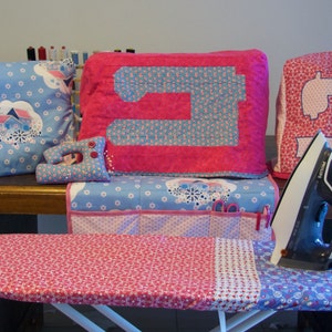 May include: A set of pink and blue sewing machine covers and a pink and blue ironing board cover. The covers are decorated with a floral pattern and a sewing machine design. There is also a black iron on the ironing board.