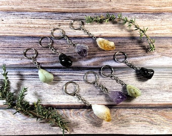 May include: A collection of keychains featuring various polished gemstones, including amethyst, citrine, and green stones. Each keychain has a silver-colored chain and ring. The gemstones are displayed on a wooden surface with sprigs of rosemary.