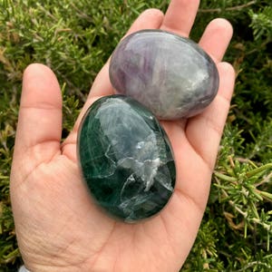 May include: Two polished fluorite stones in hand. One is a deep green color, the other is a mix of purple and gray. The stones have a smooth, rounded shape and a glossy finish. The background is blurred green foliage.