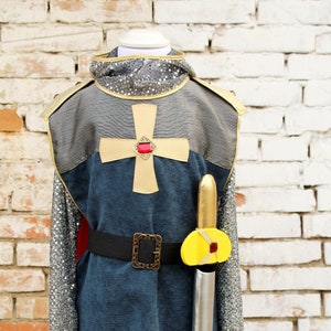 Pode incluir: Um traje de cavaleiro para crianças com uma túnica azul, mangas de cota de malha, uma sobreveste vermelha, um cinto amarelo, uma espada amarela e vermelha e uma cruz vermelha no peito.