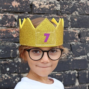 May include: A child wearing a gold glitter crown with the number 7 on it. The child is wearing black framed glasses and a white long-sleeved shirt.