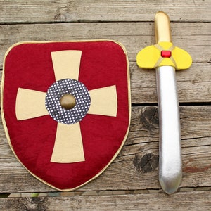 Pode incluir: Um escudo vermelho com uma cruz dourada e um círculo central de bolinhas pretas e brancas com um rebite dourado, acompanhado por uma espada prateada com cabo dourado e um pequeno detalhe retangular vermelho no centro da guarda amarela.