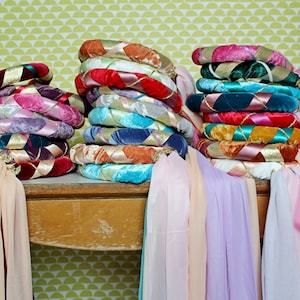 May include: Stacks of colorful, decorative headbands with velvet and satin ribbons. The headbands are in various colors, including pink, red, blue, and gold. Sheer fabric drapes down from the headbands.