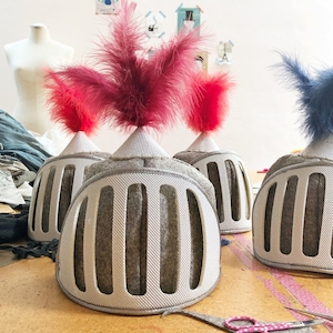 May include: Three gray felt knight helmets with red and blue feathers. The helmets have a grid pattern and are sitting on a wooden surface.