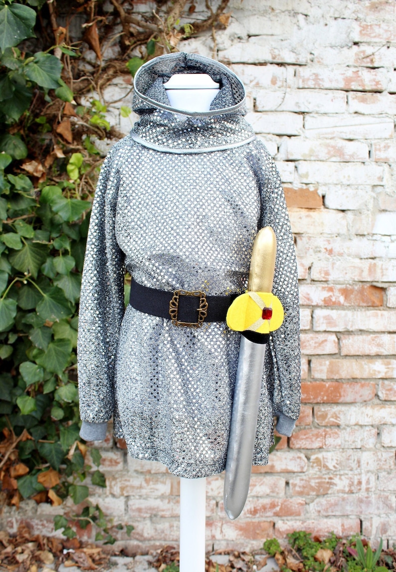 Op de afbeelding: Een zilveren pailletten tuniek met een zwarte riem en een geel en gouden speelgoedzwaard. De tuniek heeft een hoge kraag en lange mouwen.