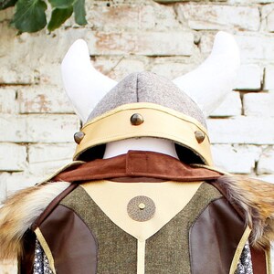 May include: A brown and green costume with a faux fur collar and a Viking helmet with white horns. The helmet has a gold band with metal accents.
