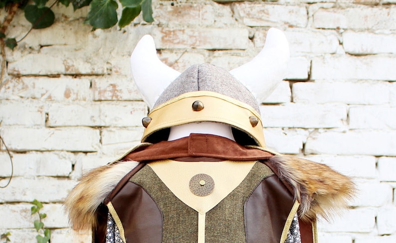 May include: A brown and green costume with a faux fur collar and a Viking helmet with white horns. The helmet has a gold band with metal accents.