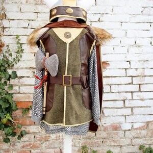 May include: A brown and green costume with a faux fur trim, a brown leather belt, and a grey axe. The costume is worn by a mannequin with a white helmet with gold accents.