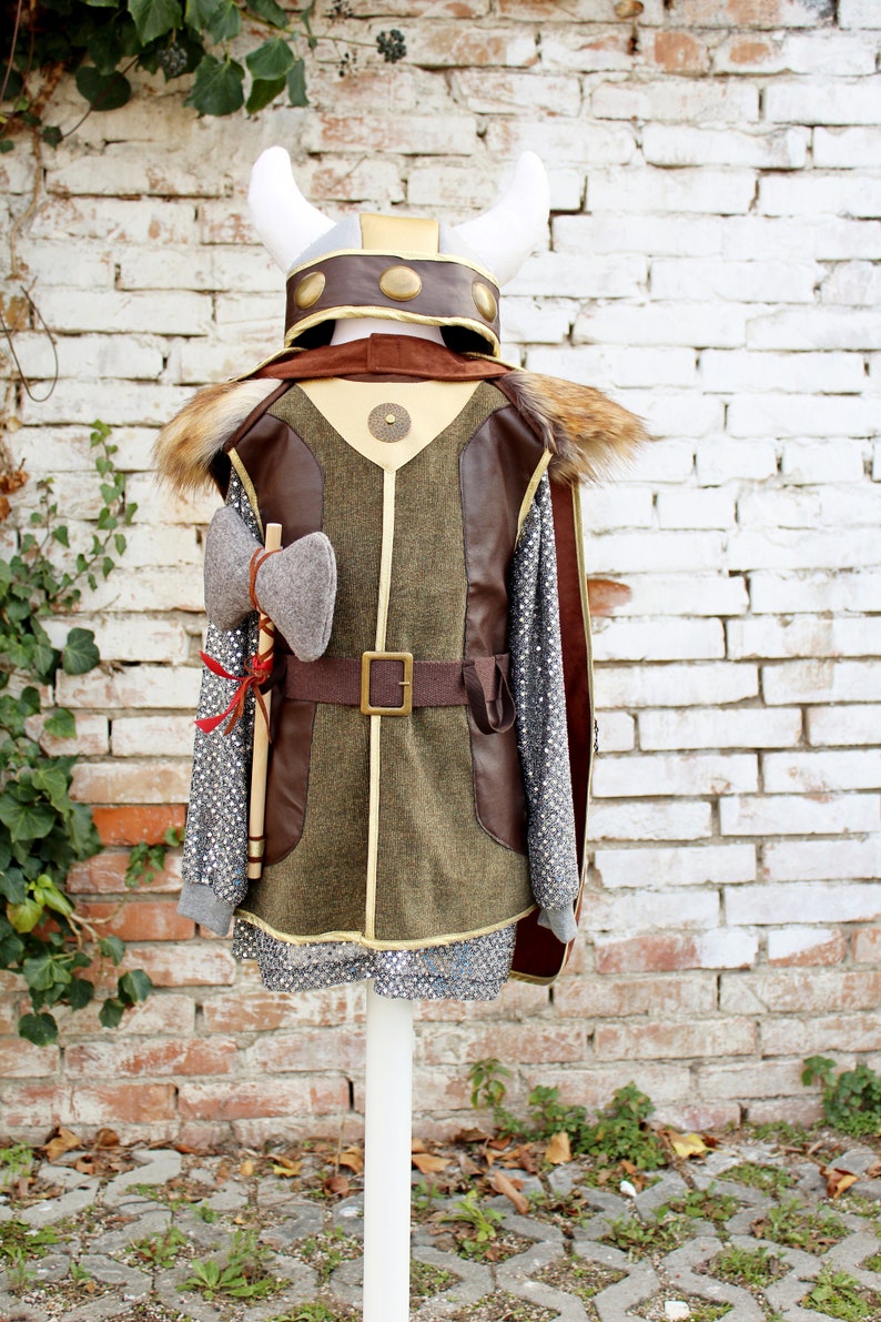 May include: A brown and green costume with a faux fur trim, a brown leather belt, and a grey axe. The costume is worn by a mannequin with a white helmet with gold accents.