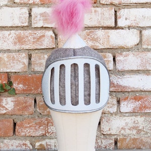 May include: A gray felt knight helmet with a pink pom-pom on top. The helmet has a silver face shield with five vertical bars.