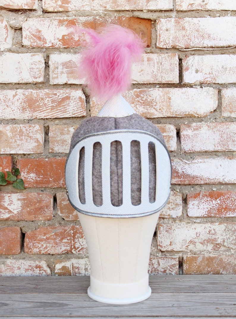 May include: A gray felt knight helmet with a pink pom-pom on top. The helmet has a silver face shield with five vertical bars.