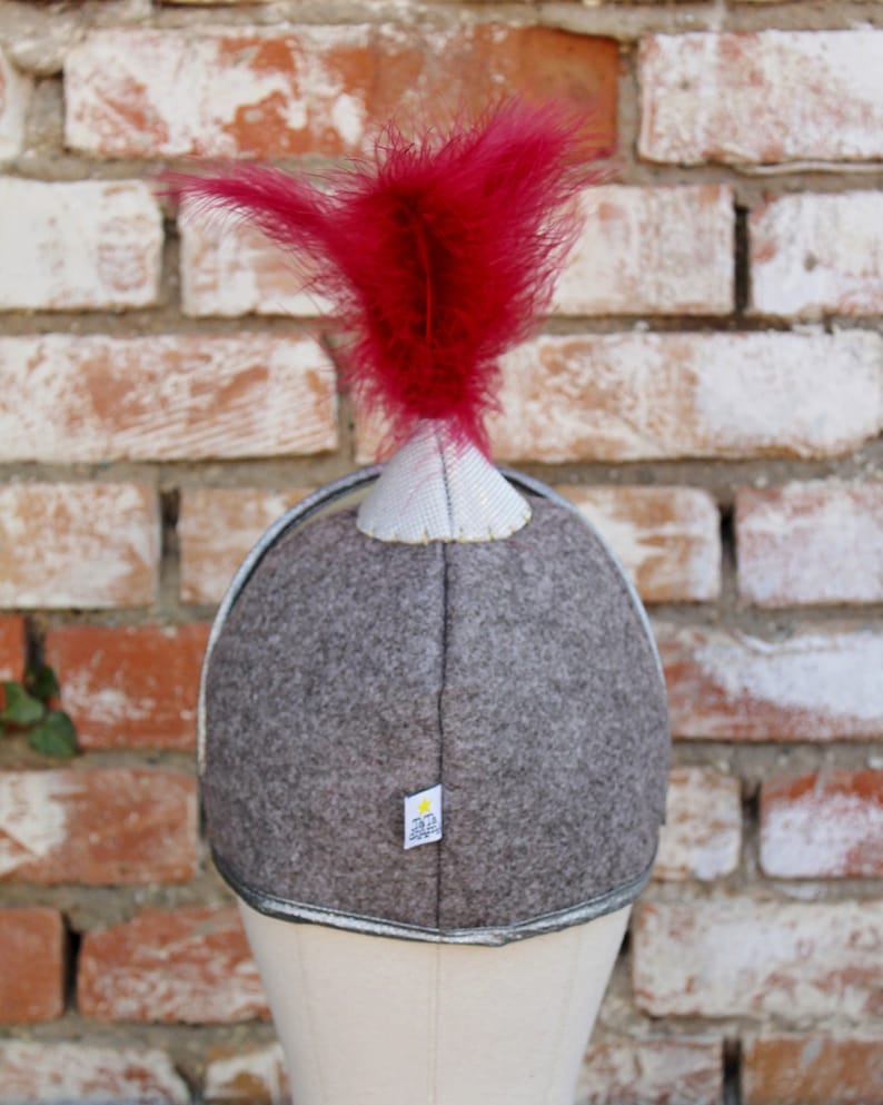 May include: A gray felt helmet with a silver trim and a red feather on top. The helmet has a small white label with the text "323" on it.