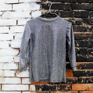 May include: A silver sequined long-sleeved shirt hanging on a white hanger against a white brick and black brick wall background.