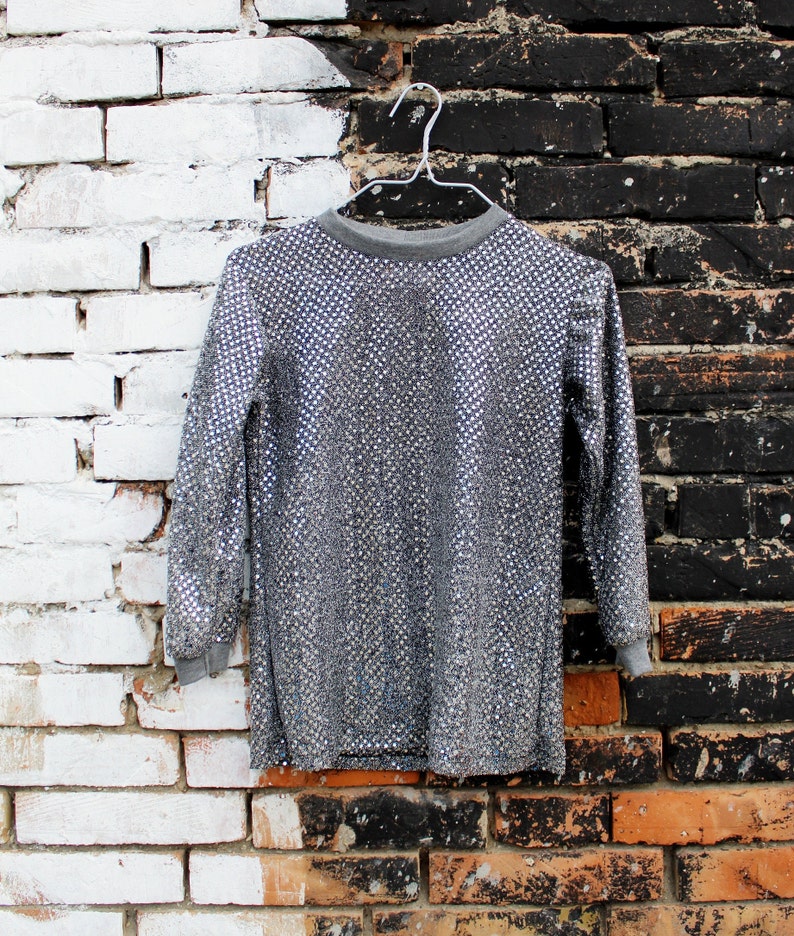 May include: A silver sequined long-sleeved shirt hanging on a white hanger against a white brick and black brick wall background.