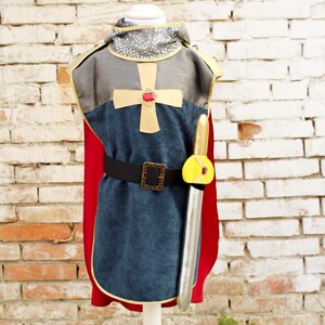 Pode incluir: Um traje de cavaleiro composto por uma t&uacute;nica azul com uma cruz dourada e uma gema vermelha, um capuz prateado estilo cota de malha, uma capa vermelha e um cinto preto com fivela dourada. Uma espada de brinquedo prateada est&aacute; presa ao cinto.
