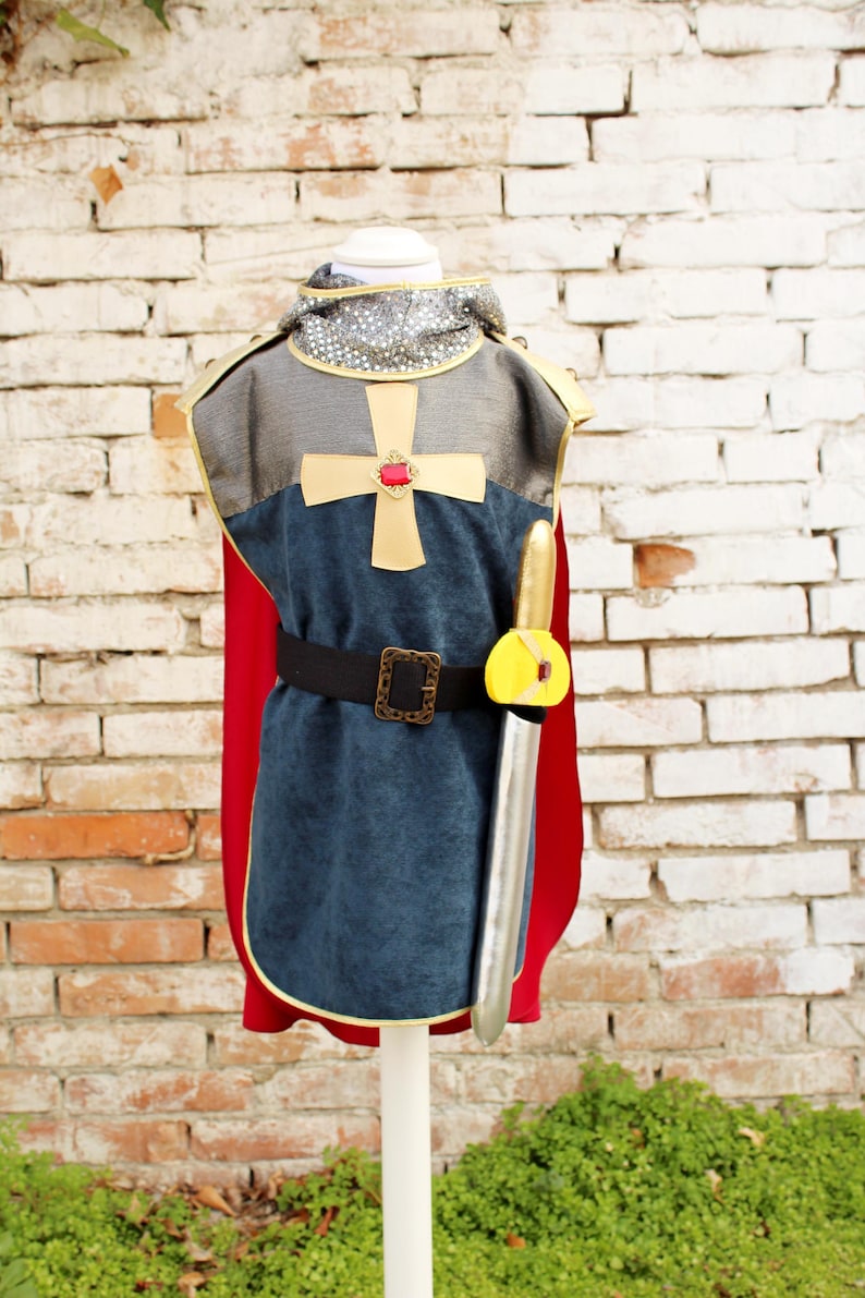 Pode incluir: Um traje de cavaleiro composto por uma t&uacute;nica azul com uma cruz dourada e uma gema vermelha, um capuz prateado estilo cota de malha, uma capa vermelha e um cinto preto com fivela dourada. Uma espada de brinquedo prateada est&aacute; presa ao cinto.
