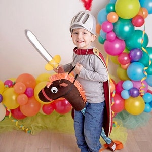 May include: A young child dressed as a knight, wearing a silver helmet with a red feather, holds a toy sword and rides a brown toy horse. The child is surrounded by colorful balloons.