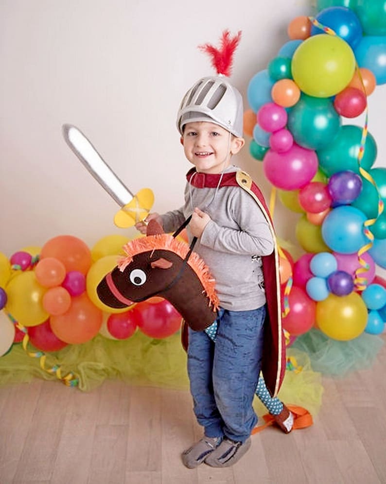 May include: A young child dressed as a knight, wearing a silver helmet with a red feather, holds a toy sword and rides a brown toy horse. The child is surrounded by colorful balloons.