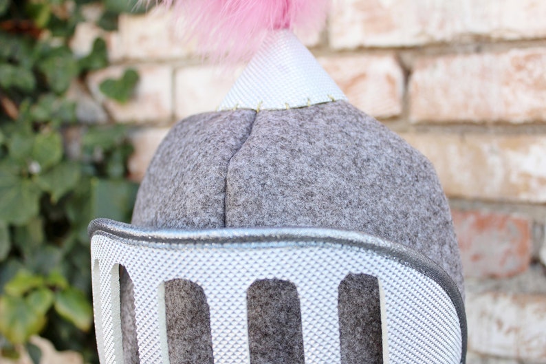 May include: A gray felt knight helmet with a silver plastic face guard and a pink feather plume on top.