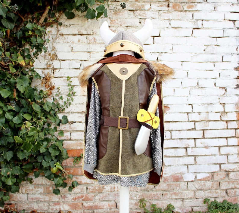 May include: A brown and green Viking costume for kids with a faux fur trim, a chainmail tunic, a brown leather belt, and a white helmet with horns. The costume includes a yellow and brown toy sword.