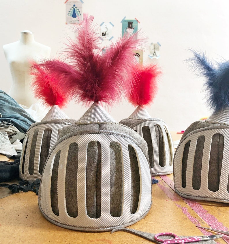 May include: Three gray felt knight helmets with red and blue feathers. The helmets have a grid pattern and are sitting on a wooden surface.