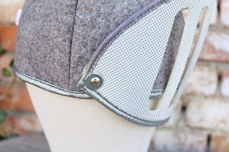 May include: A gray felt helmet with a silver trim and a silver textured panel. The helmet has a metal button on the side.