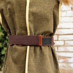 Pode incluir: Um colete sem mangas verde-oliva com debrum amarelo-claro nas bordas e um cinto de tecido marrom com uma tira de fecho de velcro preta. O colete tem uma superf&iacute;cie texturizada e um acabamento semelhante a pelo marrom na gola.