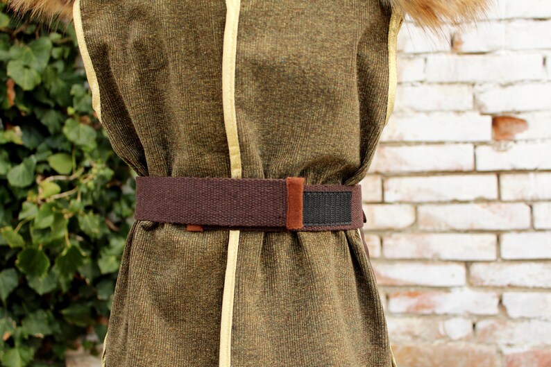 Pode incluir: Um colete sem mangas verde-oliva com debrum amarelo-claro nas bordas e um cinto de tecido marrom com uma tira de fecho de velcro preta. O colete tem uma superf&iacute;cie texturizada e um acabamento semelhante a pelo marrom na gola.