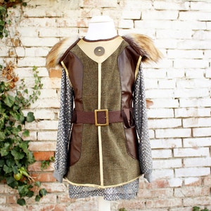 May include: A brown and green faux leather vest with a fur collar and a brown belt with a metal buckle. The vest has a decorative metal piece on the front.