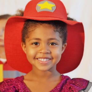 May include: A young girl wearing a red felt hat with a yellow star on top. She is smiling and wearing a sparkly purple dress.