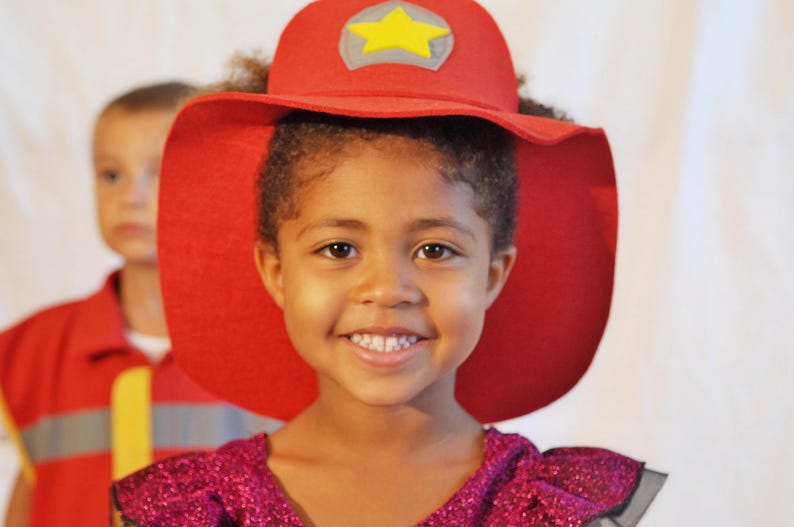 May include: A young girl wearing a red felt hat with a yellow star on top. She is smiling and wearing a sparkly purple dress.
