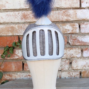 May include: A white knight helmet with a blue feather on top. The helmet has a silver faceplate with a series of vertical bars.