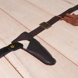 May include: A dark brown fabric belt featuring a brass-colored metal buckle, a black sheath containing a small knife with a gold star emblem on the handle, and a small brown drawstring pouch attached to the belt strap.