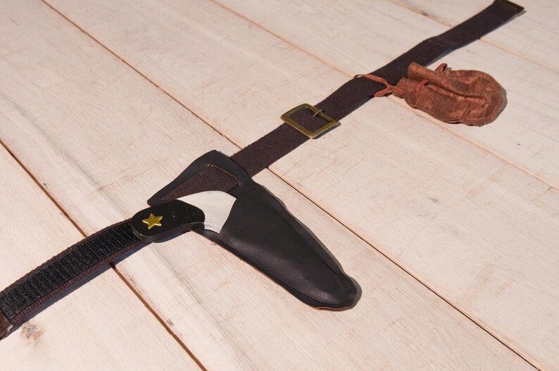 May include: A dark brown fabric belt featuring a brass-colored metal buckle, a black sheath containing a small knife with a gold star emblem on the handle, and a small brown drawstring pouch attached to the belt strap.