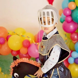 May include: A young child dressed as a knight in a silver helmet with a red feather, riding a black toy horse with orange mane. The child is surrounded by colorful balloons.