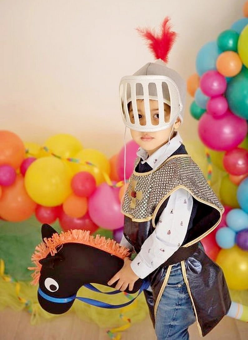 May include: A young child dressed as a knight in a silver helmet with a red feather, riding a black toy horse with orange mane. The child is surrounded by colorful balloons.