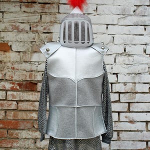 Puede incluir: Armadura de cota de malla plateada con un casco plateado con una pluma roja en la parte superior. La armadura est&aacute; en un maniqu&iacute; contra una pared de ladrillo.