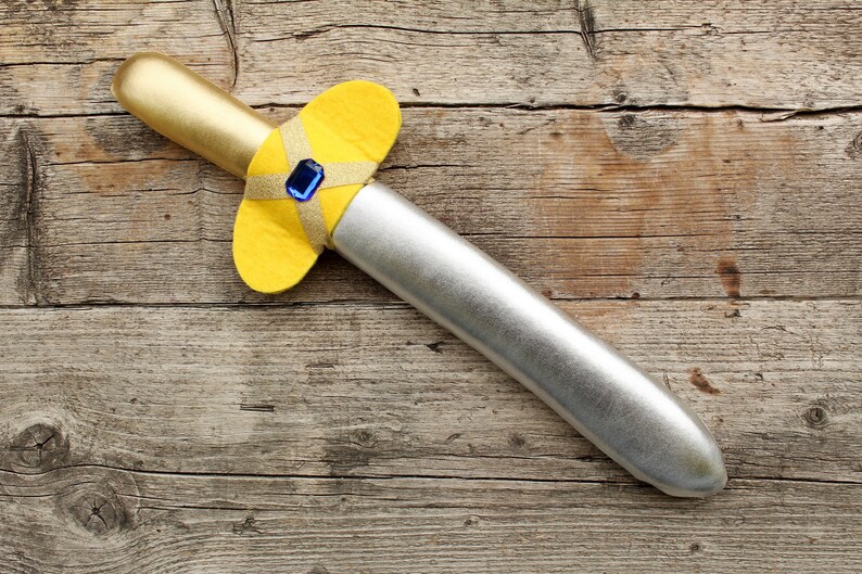 Op de afbeelding: Een zilveren en gouden speelgoedzwaard met een gele viltgreep en een blauwe edelsteen. Het zwaard ligt op een houten oppervlak.