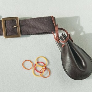 May include: A dark brown canvas belt with a rectangular brass-toned metal buckle, featuring a small dark brown leather pouch attached by a thin leather cord, accompanied by four small circular rubber bands in yellow and orange colors.