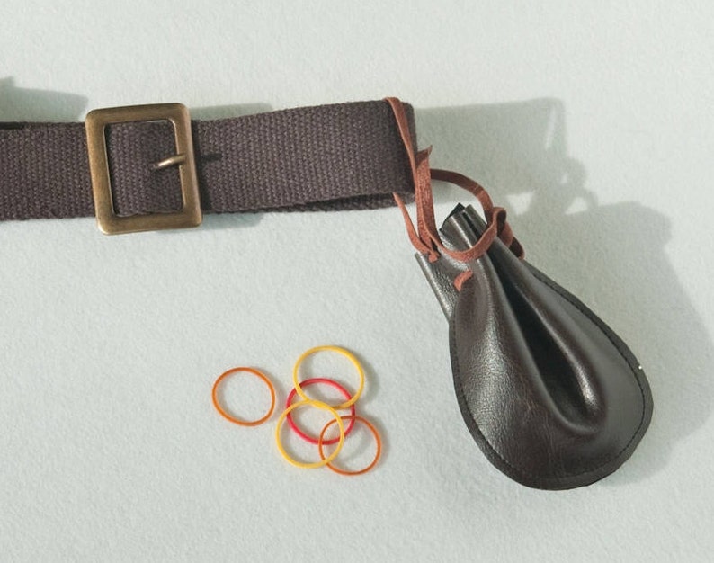 May include: A dark brown canvas belt with a rectangular brass-toned metal buckle, featuring a small dark brown leather pouch attached by a thin leather cord, accompanied by four small circular rubber bands in yellow and orange colors.