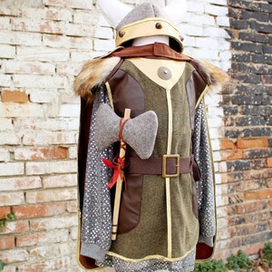 May include: A child's costume of a Viking warrior with a brown leather and green fabric tunic, a gray fur-trimmed cape, a white helmet with gold accents, and a gray felt axe.