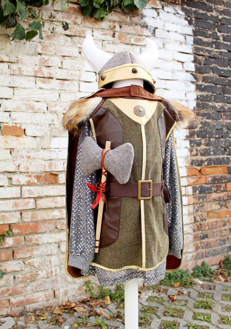 May include: A child's costume of a Viking warrior with a brown leather and green fabric tunic, a gray fur-trimmed cape, a white helmet with gold accents, and a gray felt axe.