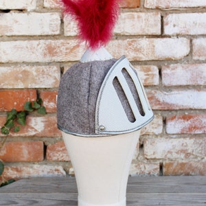 Puede incluir: Un casco de caballero gris y blanco con una pluma roja en la parte superior. El casco est&aacute; en una cabeza de maniqu&iacute; blanca.