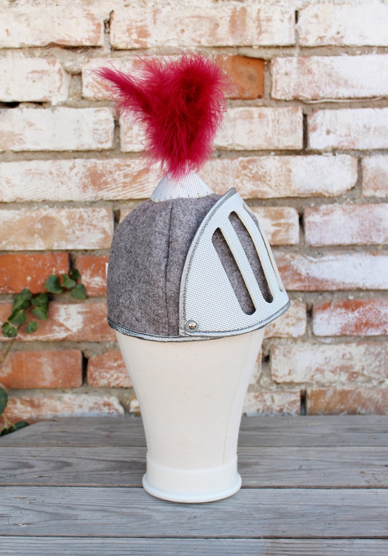 Puede incluir: Un casco de caballero gris y blanco con una pluma roja en la parte superior. El casco est&aacute; en una cabeza de maniqu&iacute; blanca.