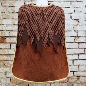 May include: Brown hooded cape with a mesh top and a gold trim. The cape is on a white mannequin against a brick wall background.