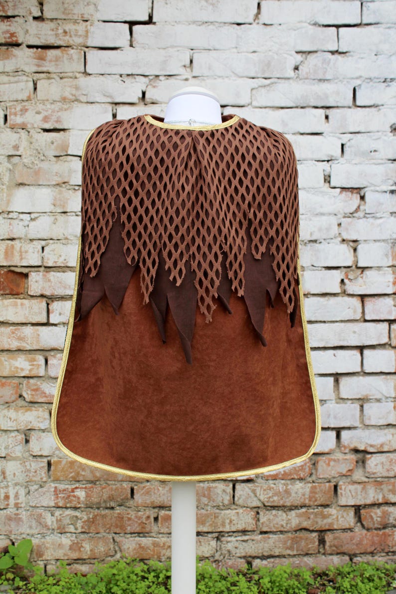 May include: Brown hooded cape with a mesh top and a gold trim. The cape is on a white mannequin against a brick wall background.